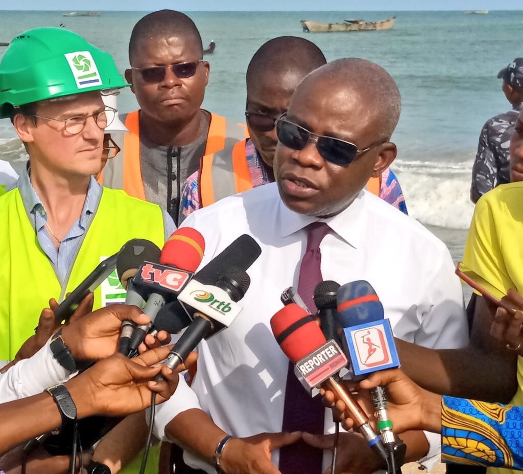 Un homme en chemise blanche et cravate sombre répond aux questions des journalistes sur une plage avec plusieurs personnes en arrière-plan.