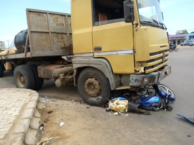 Camion jaune immobilisé avec une moto bleu écrasée sous la roue avant droite sur une route urbaine