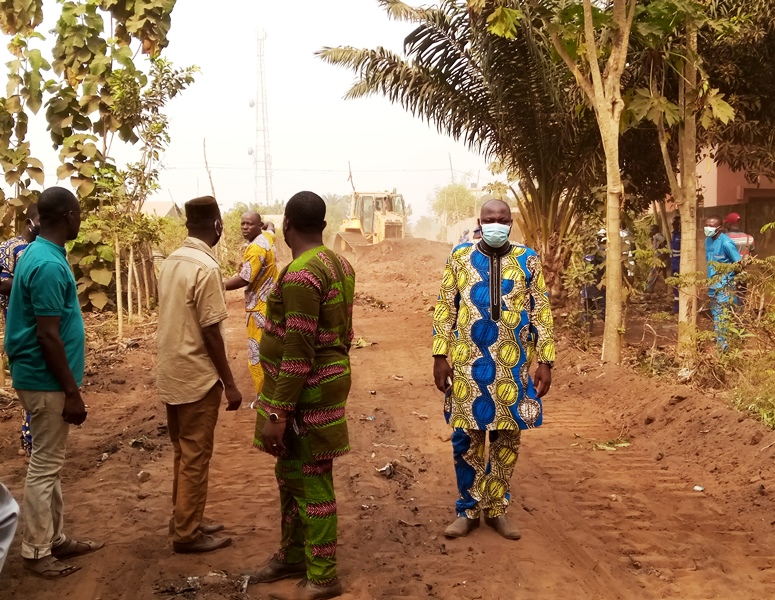 Un groupe d’hommes debout sur un chemin en terre, un engin de chantier en arrière-plan soulève de la poussière