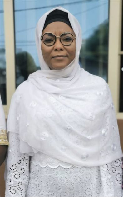Femme portant un voile blanc orné de broderies et des lunettes rondes pose devant une façade vitrée