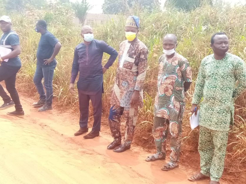 Six hommes debout le long d’un chemin de terre herbeux, trois portent des masques et deux tiennent des papiers