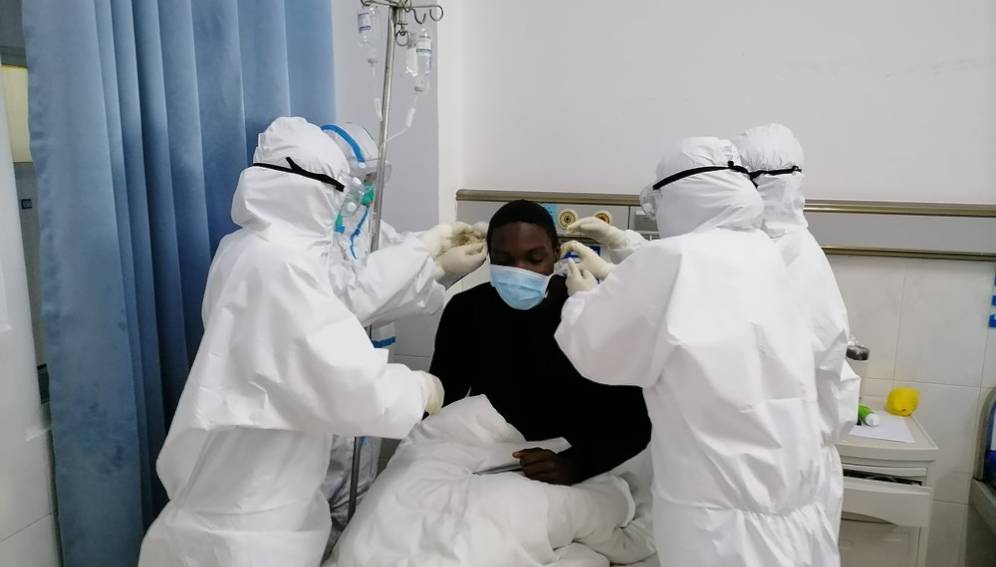 Deux soignants en tenue de protection complète entourent un patient masqué allongé dans un lit d'hôpital