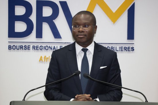 Homme en costume sombre et cravate debout derrière un pupitre avec microphones devant un logo BRVM sur un mur blanc