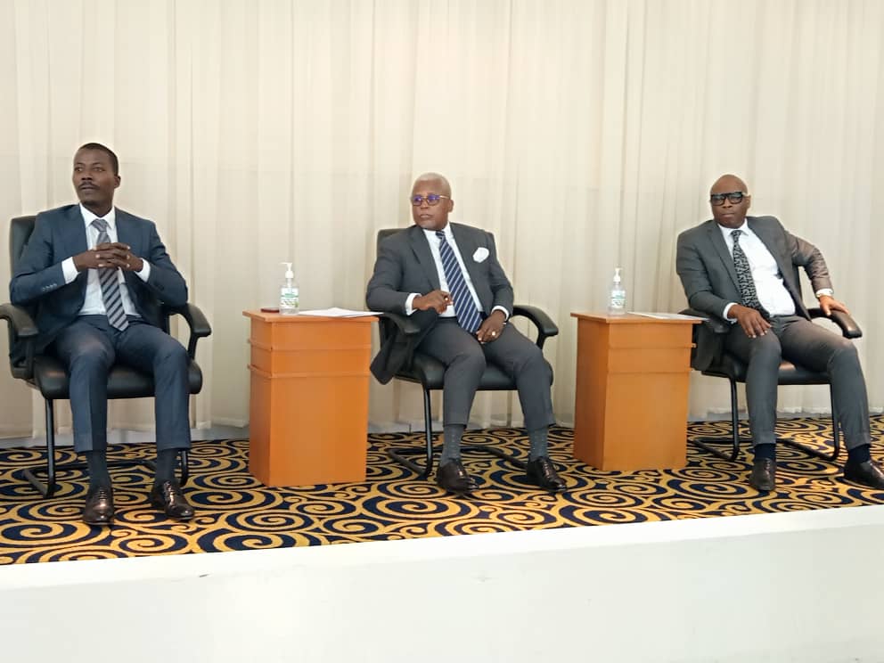 Trois hommes en costume assis sur des fauteuils dans une salle avec tapis à motifs et murs clairs