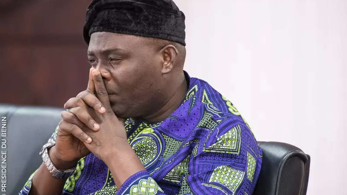 Un homme en tenue traditionnelle africaine à motifs verts et bleus, la tête appuyée sur les mains jointes, regard pensif sur fond clair
