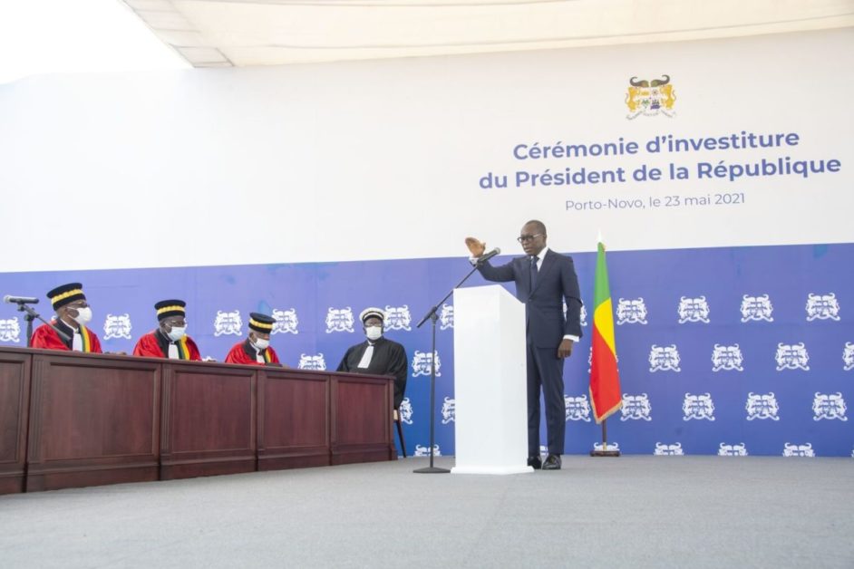 Un homme en costume prononce un discours lors d’une cérémonie avec plusieurs personnes en toges assises à une table, un drapeau est visible.