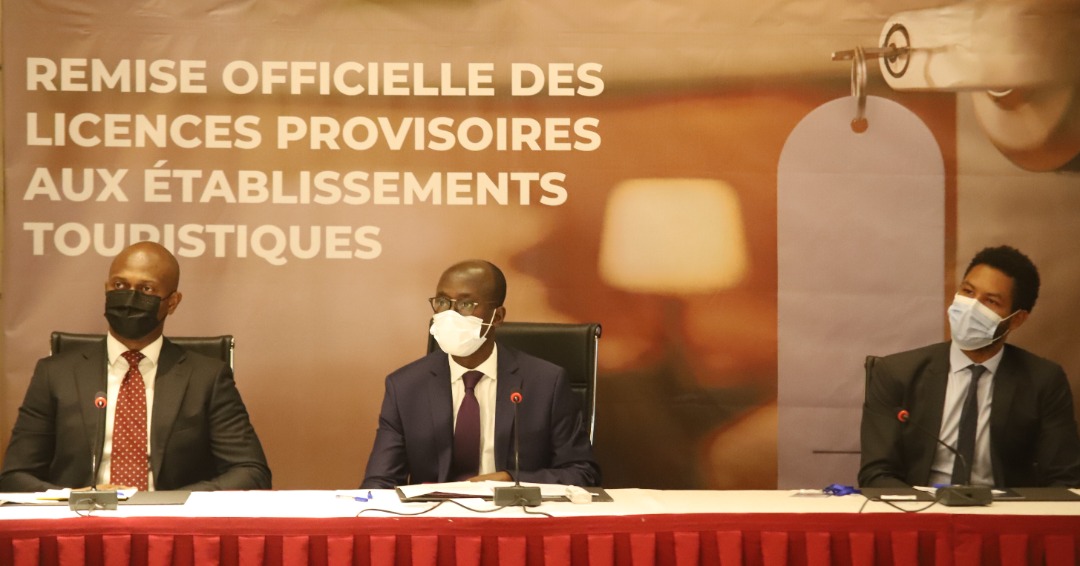 Trois hommes en costume et masque sont assis à une table devant une banderole annonçant des licences provisoires pour établissements touristiques