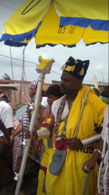 Un homme en habits traditionnels jaunes et or tenant un sceptre sous un grand parasol jaune entouré de plusieurs personnes
