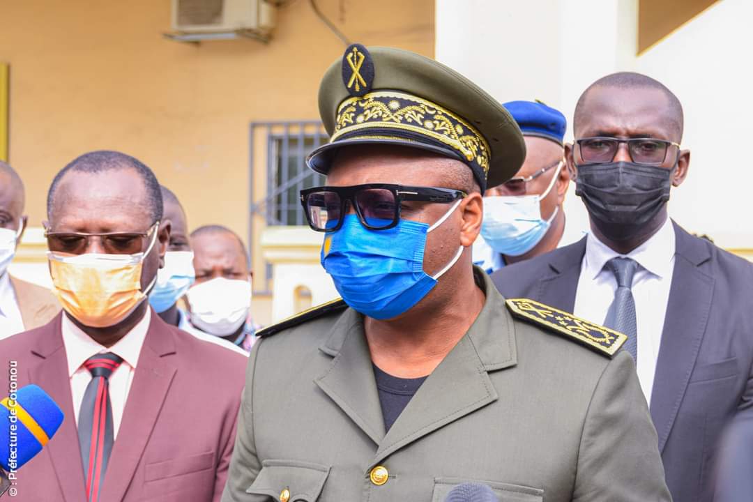 Un homme en uniforme militaire et lunettes noires parle devant plusieurs personnes portant des masques chirurgicaux