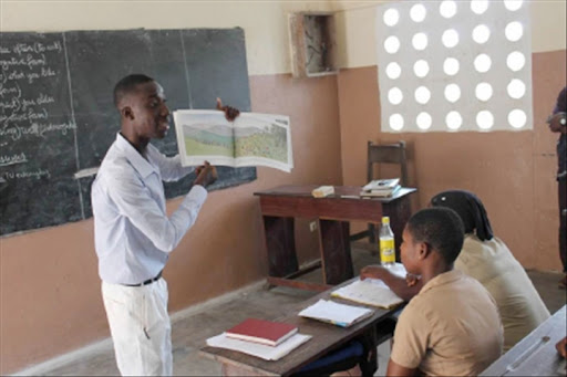 Un enseignant montre une image à deux élèves assis dans une salle de classe simple et éclairée par une fenêtre à trous circulaires