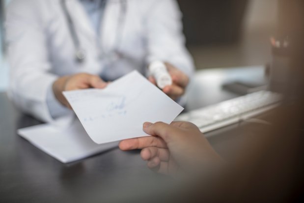 Une personne tend un document tenu par une autre personne en blouse blanche dans un bureau flou en arrière-plan