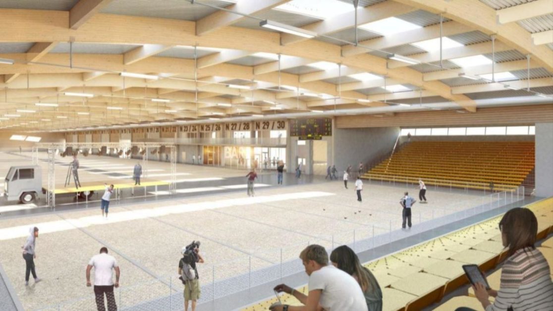 Intérieur d’un boulodrome avec terrain de pétanque, gradins jaunes et plusieurs personnes en activité et spectateurs assis