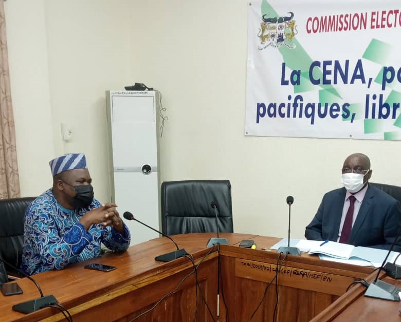 Deux hommes masqués discutent assis face à face autour d’une table dans une salle de réunion officielle