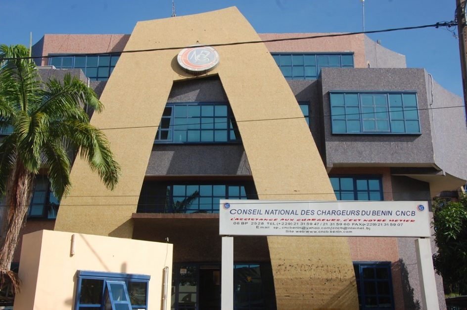 Façade d un bâtiment administratif beige avec un panneau blanc portant un nom officiel et une palme à gauche