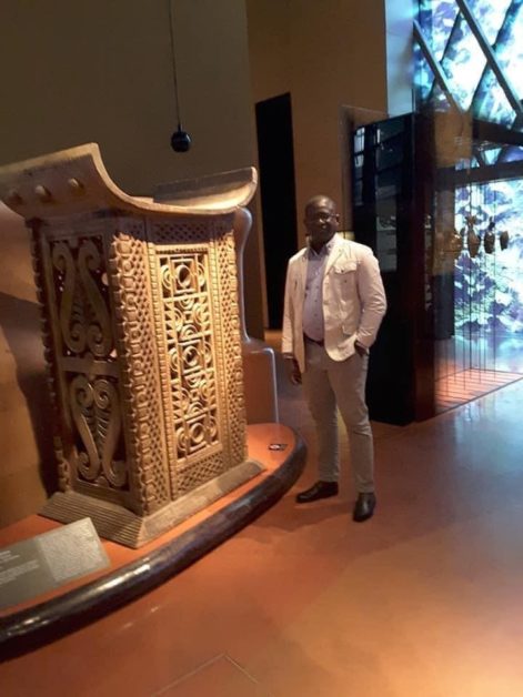 Un homme en costume clair pose près d une grande sculpture ou objet décoratif en bois sculpté dans une salle d exposition lumineuse