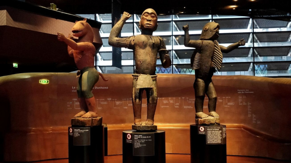Trois statues anthropomorphes en bois exposées sur des socles dans un musée éclairé naturellement par une large baie vitrée