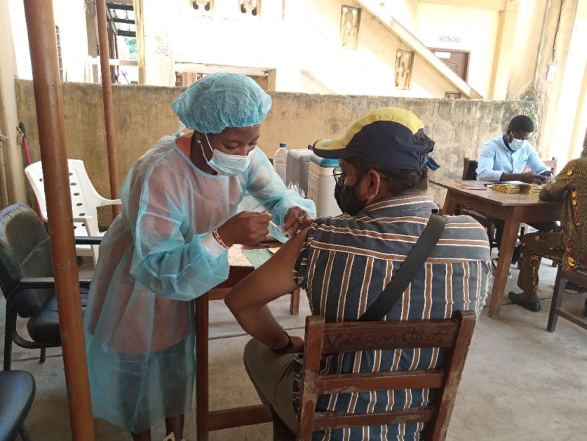 Un soignant en tenue de protection administre un vaccin à un homme assis portant un masque et une casquette dans un local simple