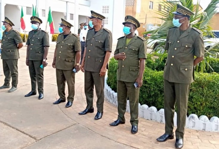 Sept hommes en uniforme et masque sanitaire debout alignés dans un espace extérieur près d’un bâtiment et des drapeaux colorés