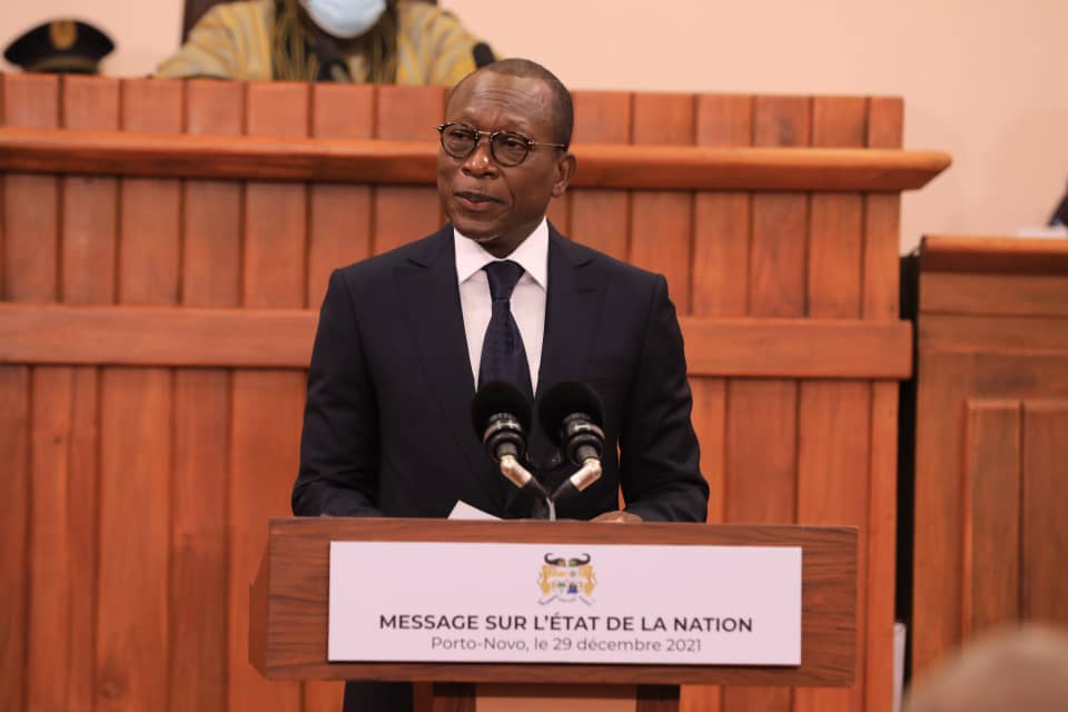 Un homme en costume sombre et lunettes parle derrière un pupitre avec un panneau indiquant un message sur l’état de la nation