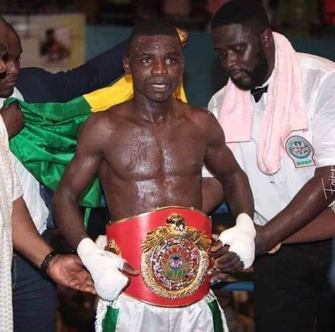Un boxeur torse nu porte une ceinture de champion assisté par deux hommes dans un environnement sportif.