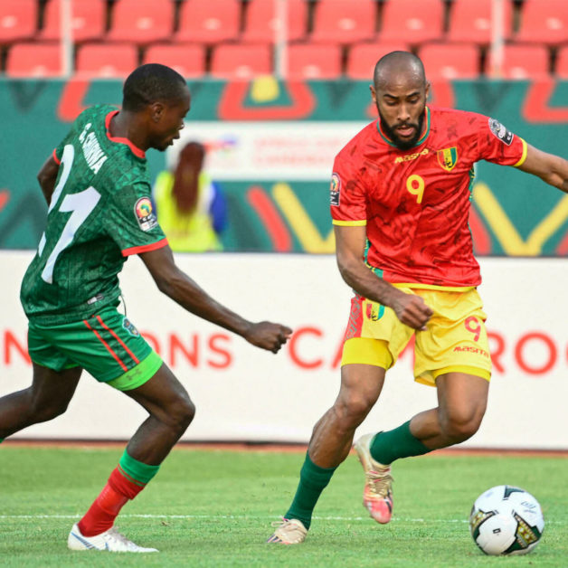 Deux joueurs de football en action sur le terrain, l'un en maillot rouge et jaune dribblant le ballon face à un adversaire en vert et rouge