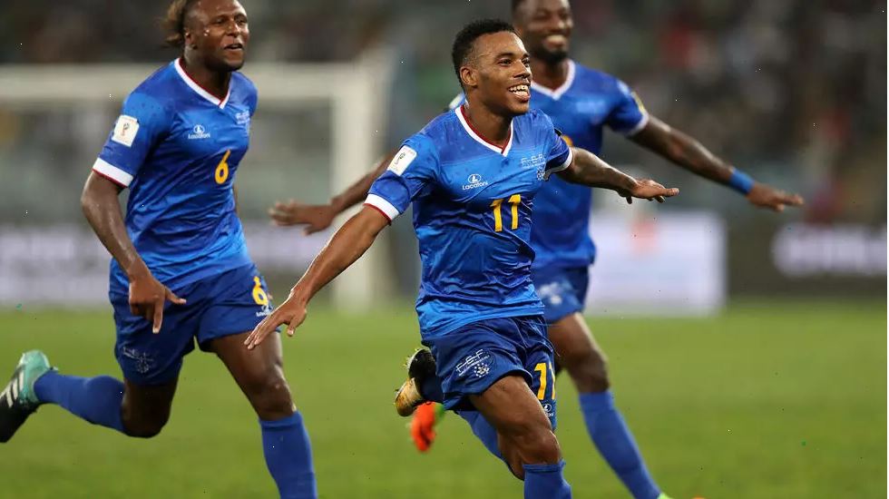 Trois joueurs de football en tenue bleue célèbrent un but sur le terrain durant un match nocturne