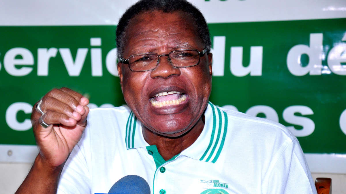Un homme portant des lunettes et un polo blanc s'exprime vivement devant un micro avec un panneau vert en arrière-plan