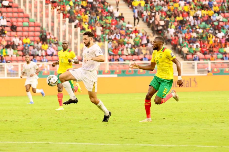 Un joueur en maillot blanc contrôle le ballon face à un adversaire en maillot jaune et vert sur un terrain de football devant des tribunes partiellement remplie