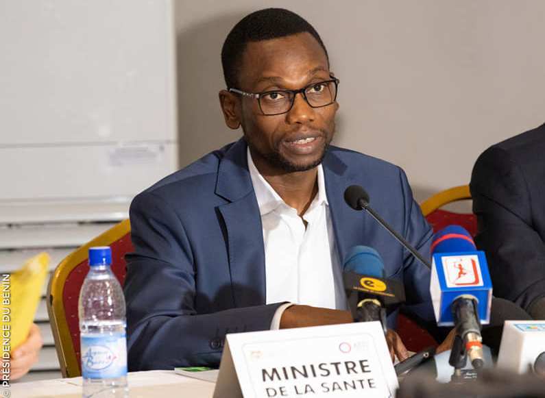 Un homme en costume bleu parle devant plusieurs micros lors d’une réunion officielle avec un panneau indiquant « Ministre de la Santé