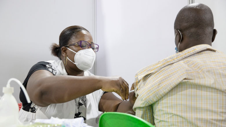 Une femme en blouse et masque vaccine un homme vu de dos assis sur une chaise verte dans un centre de vaccination