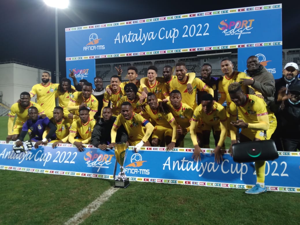Une équipe de football en maillots jaunes pose ensemble derrière un panneau Antalya Cup 2022 avec un trophée devant eux sur un terrain de nuit