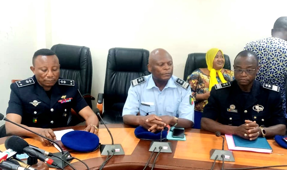Trois hommes en uniforme assis à une table de conférence avec des casquettes posées devant eux et une femme souriante en arrière-plan