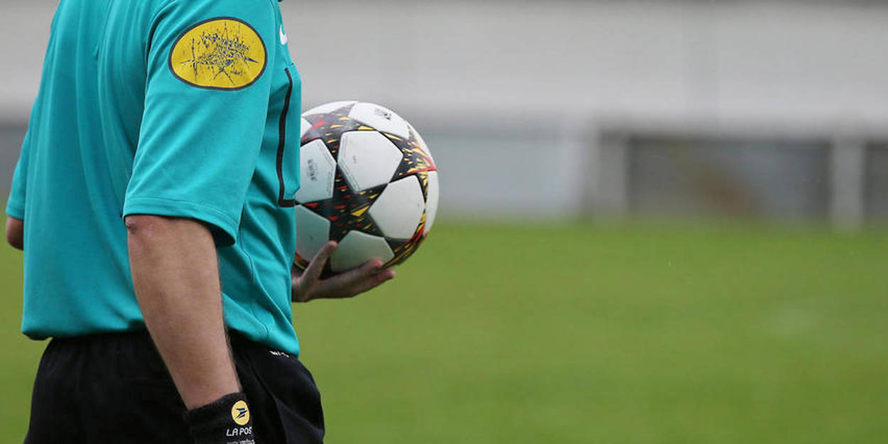 Arbitre en tenue turquoise tenant un ballon de football noir et blanc sur un terrain vert