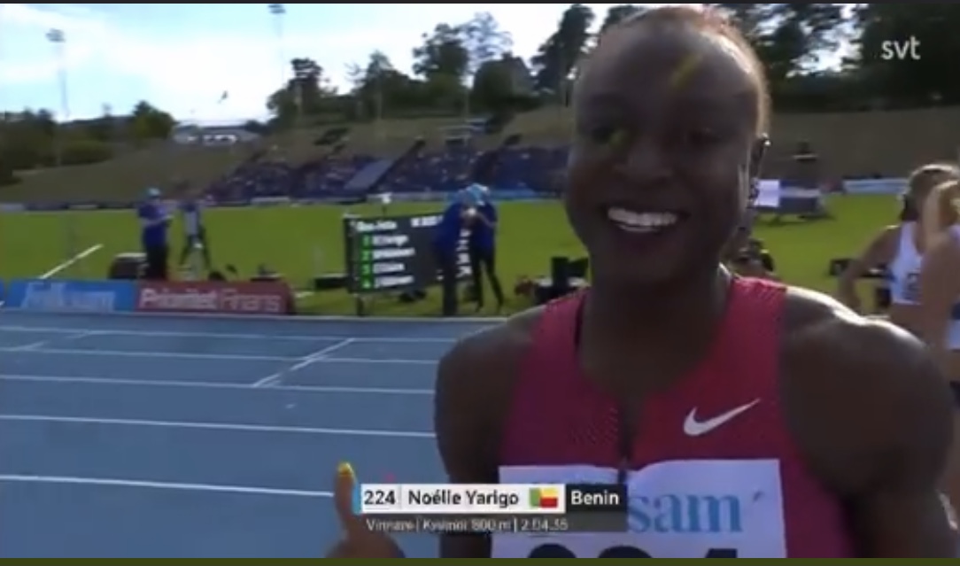 Femme souriante en tenue de course rouge sur piste d’athlétisme bleue avec gradins et écran en arrière-plan