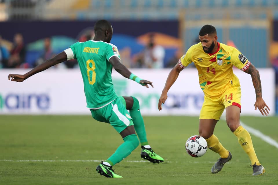 Un joueur en maillot jaune contrôle le ballon face à un adversaire en vert sur un terrain de football
