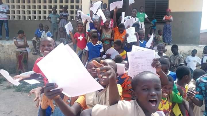 Enfants rassemblés devant un bâtiment, brandissant des feuilles avec enthousiasme et sourires nombreux
