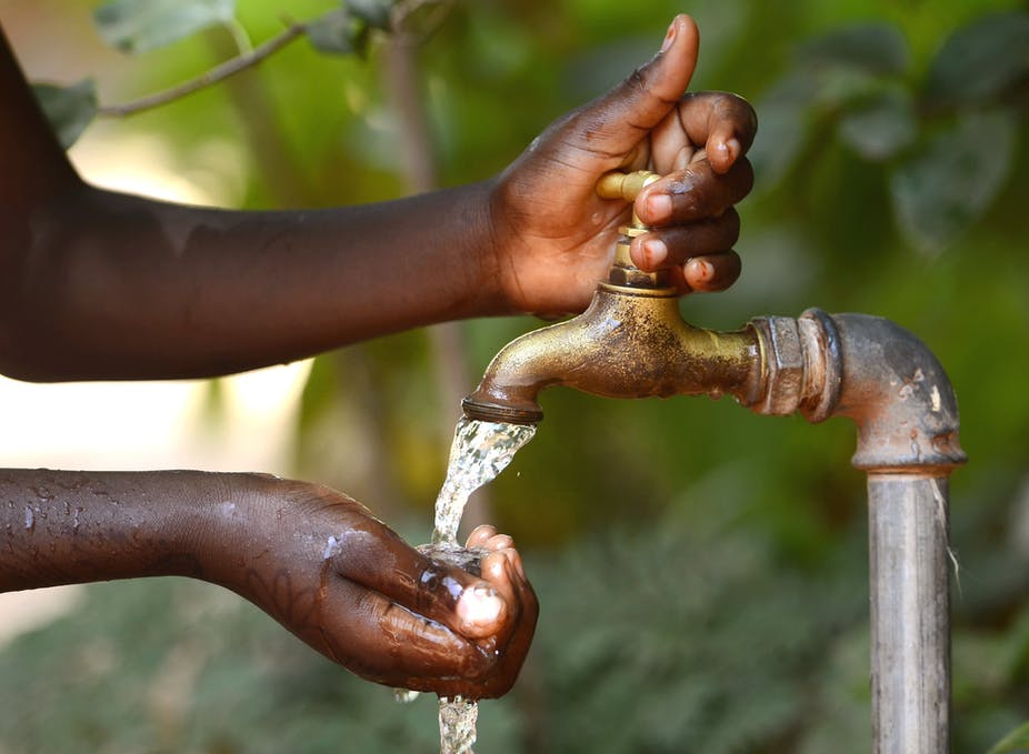 Mains noires recueillant de l’eau claire s’écoulant d’un robinet en extérieur avec un arrière-plan flou vert