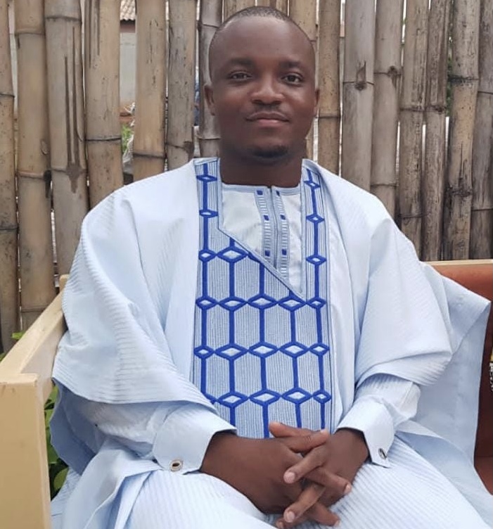 Homme assis en tenue traditionnelle blanche ornée de motifs bleus devant un mur en bambou