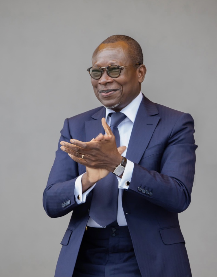 Un homme en costume bleu marine et lunettes sourit en applaudissant devant un fond gris uniforme