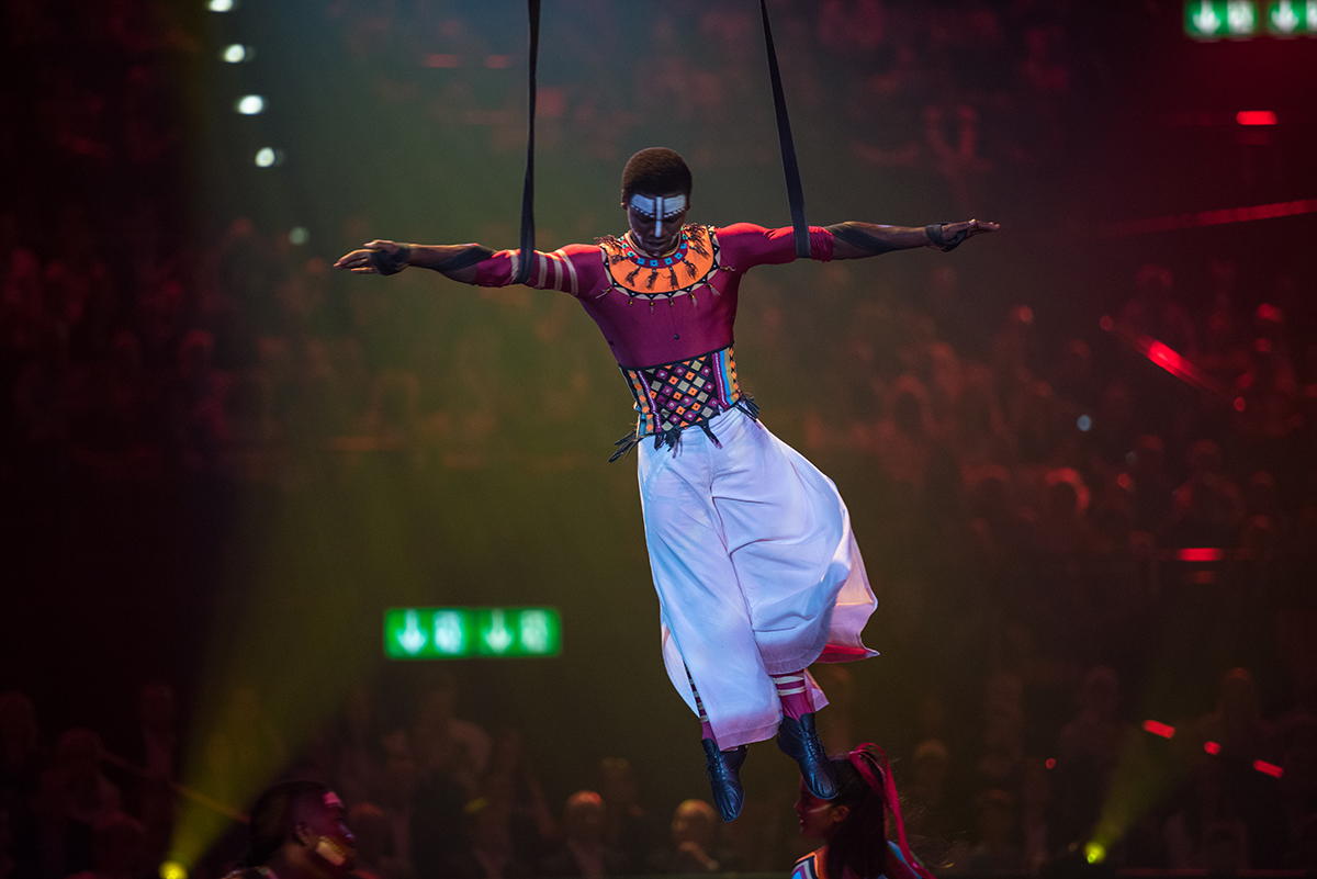 Un artiste vêtu d’un costume coloré et maquillé, suspendu dans les airs lors d’un spectacle devant un public nombreux