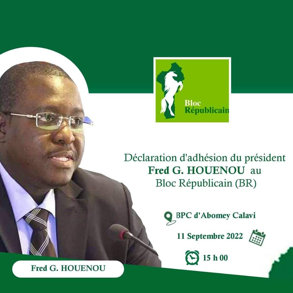 Homme en costume sombre et lunettes parlant devant un micro avec un logo vert Bloc Républicain en arrière-plan