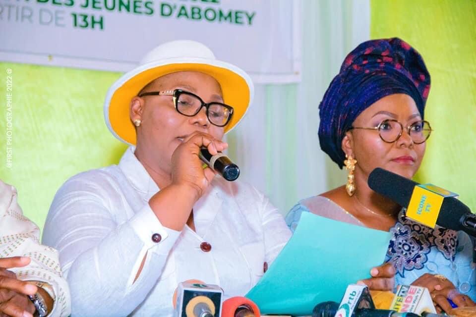 Deux femmes assises, l’une en chemise blanche et chapeau clair parle dans un micro, l’autre en tenue traditionnelle bleue écoute avec un micro devant elle
