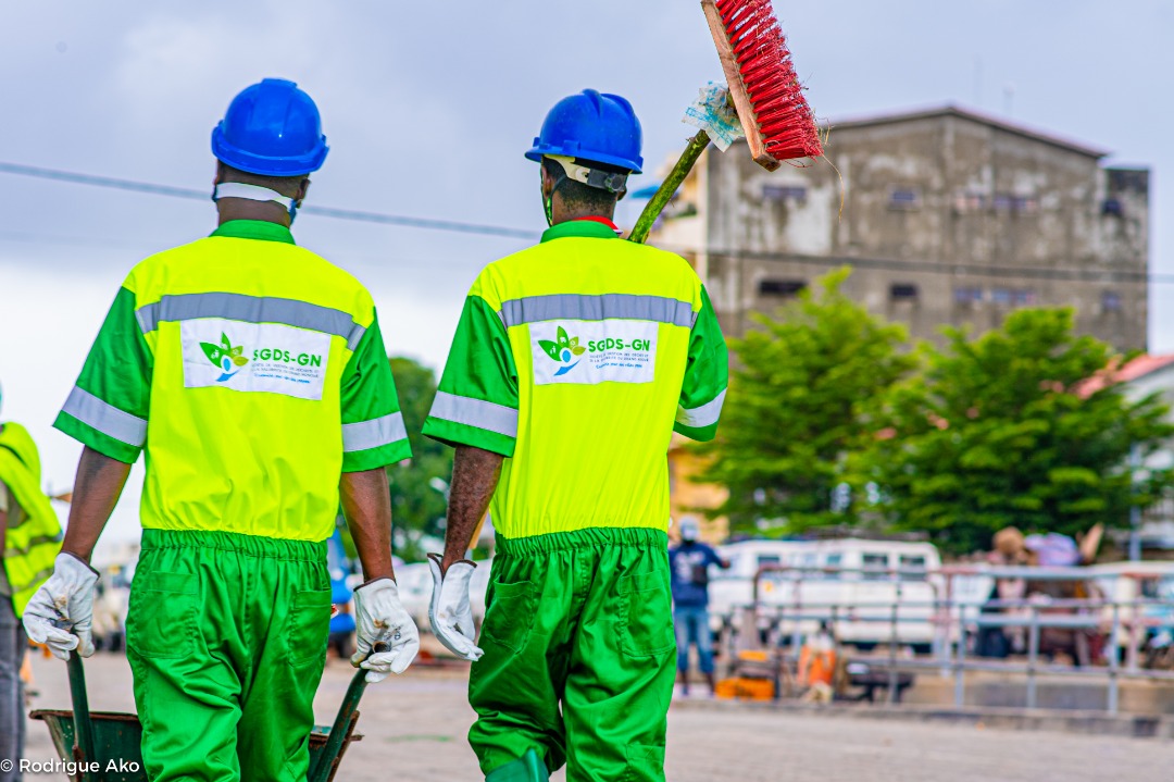 Deux employés en uniforme vert et jaune portent un balai et un matériel de nettoyage en extérieur