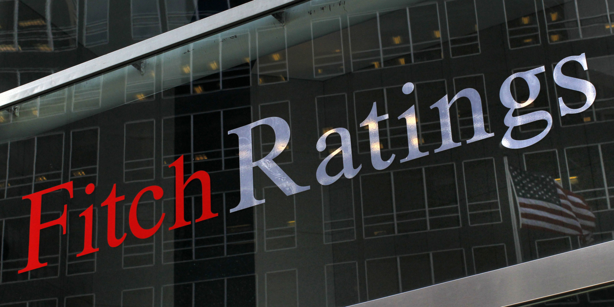 Logo Fitch Ratings en lettres rouges et blanches sur une façade en verre avec réflexion d'immeubles et un drapeau américain