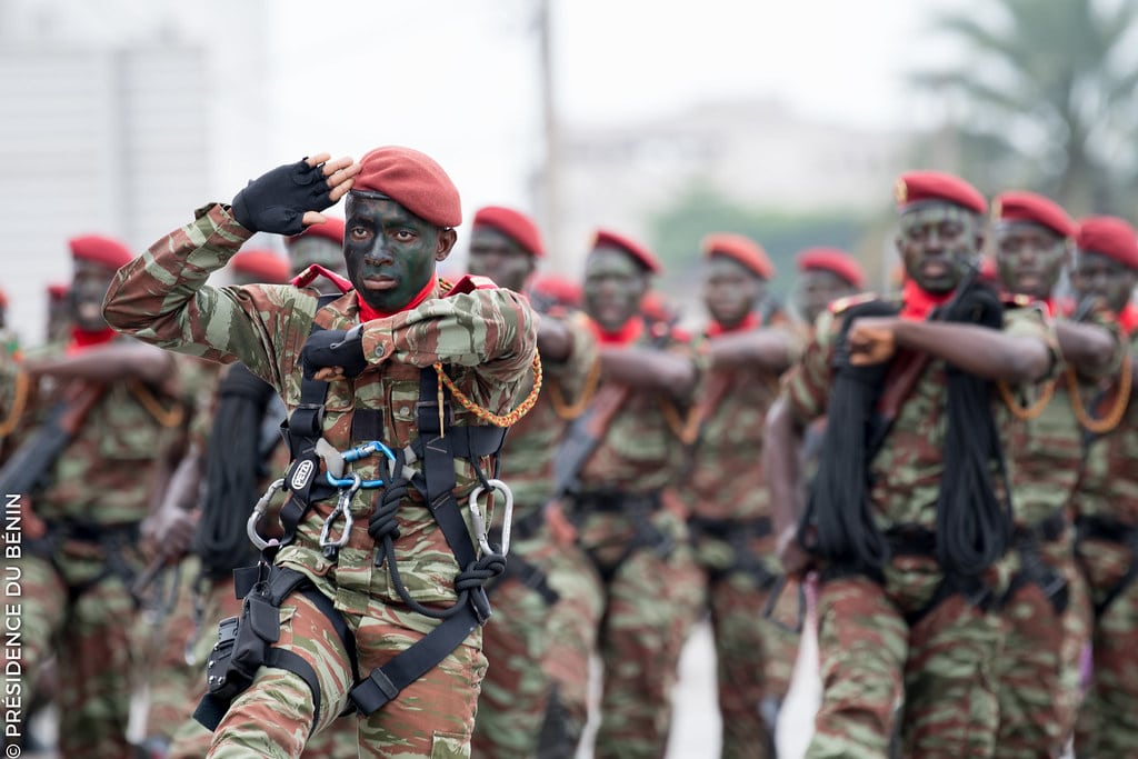 Soldats en uniforme camouflage et bérets rouges avec visage peint effectuant un salut militaire en formation.