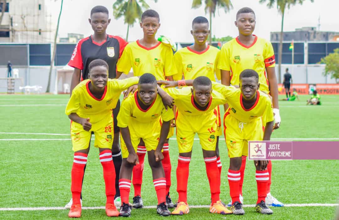 Huit jeunes joueuses en tenue de football jaune et rouge posent sur un terrain de football gazonné devant un but