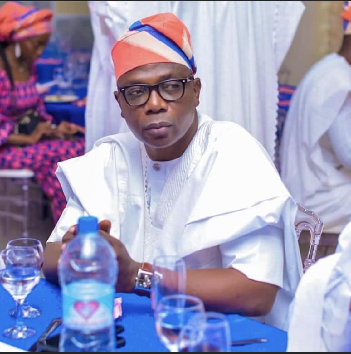 Homme portant un boubou blanc et un chapeau coloré assis à une table avec des verres et une bouteille d'eau devant lui