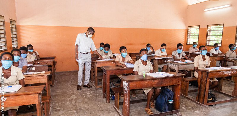 Des élèves portant uniforme et masque sont assis dans une salle de classe avec un enseignant debout parmi eux