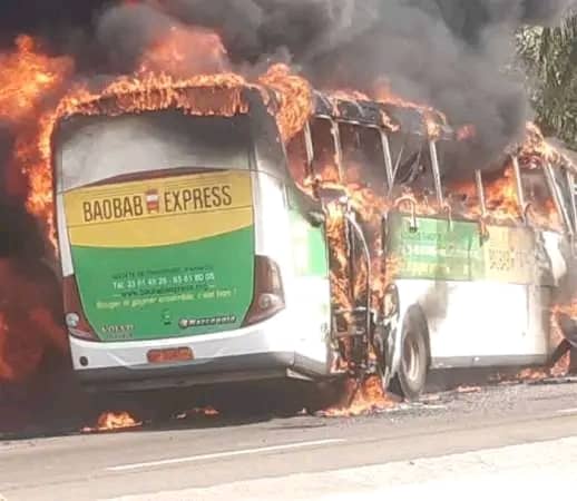 Un bus de transport est entièrement enflammé avec des flammes et de la fumée noire s’échappant du véhicule sur une route.
