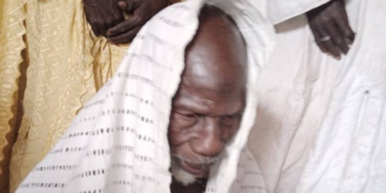 Homme âgé en tenue traditionnelle claire portant un tissu sur la tête entouré de personnes habillées en blanc